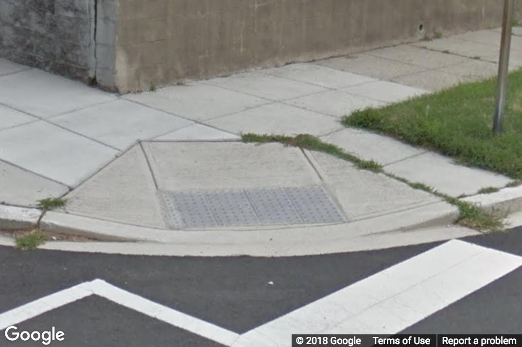 A Street View image of a curb ramp with grass growing on an edge