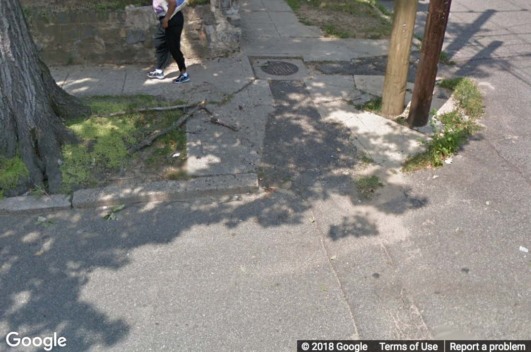 A Street View image of a curb ramp with cracks and asphalt in the center